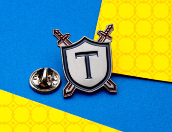 Shield-shaped lapel pin with the letter T and crossed swords behind it, shown next to a butterfly clutch backing on a blue and yellow background.