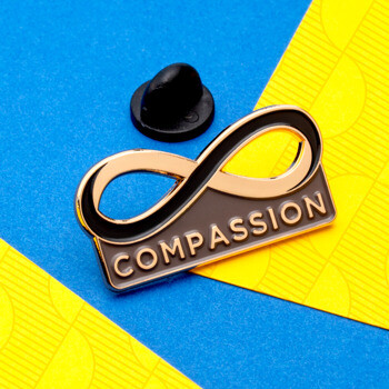 Custom lapel pin featuring a gold-toned infinity symbol and the word COMPASSION in gold text on a black enamel background.