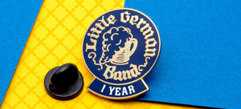 Little German Band 1 Year circular enamel lapel pin with a beer stein illustration and black rubber clutch on a blue and yellow background.