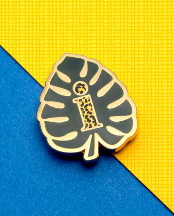 Monstera leaf lapel pin in gold and black enamel featuring a patterned letter i in the center, displayed on a blue and yellow background.